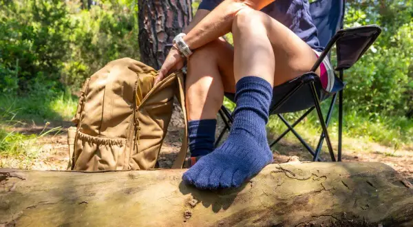 Une personne assise sur une chaise de camping, avec un sac à dos à côté. Elle porte des chaussettes bleues séparant les orteils, idéales pour la randonnée.