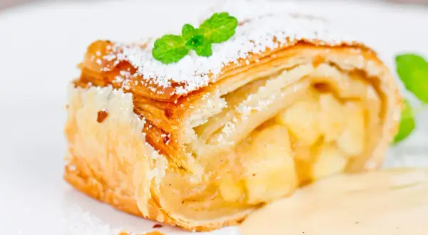 Ein Stück Apfelstrudel mit Puderzucker und Minze, serviert mit Vanillesauce. Perfekt für ein Apfelstrudel Rezept!