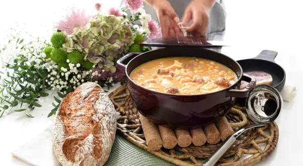 Un reconfortante plato de callos a la gallega humea en una cazuela, acompañado de pan rústico y un ramo de flores.