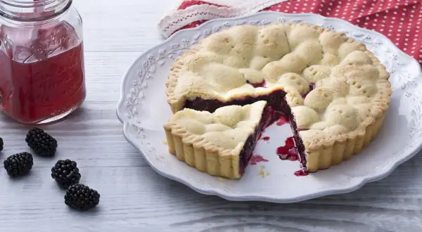 Une délicieuse tarte aux mûres, avec une part coupée, sur une assiette ornée. Des mûres fraîches et un verre de jus rouge complètent la scène.
