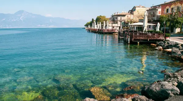 Lungolago di Gardone Riviera: cosa vedere? Case colorate, ristoranti con tavoli all'aperto e il lago cristallino.