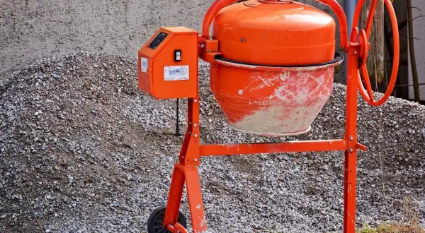 Pomarańczowa betoniarka czeka na materiały, by pokazać, jak zrobić beton. Obok stos kruszywa.