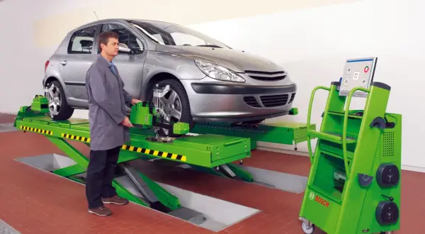 Mechanik przy samochodzie na podnośniku, obok zielona maszyna z monitorem. To profesjonalna tabelą zbieżności kół.