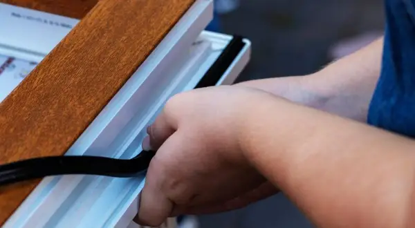 Pose d'un joint d'étanchéité pour comment insonoriser une porte. Les mains insèrent un joint noir dans le cadre blanc d'une porte imitation bois.