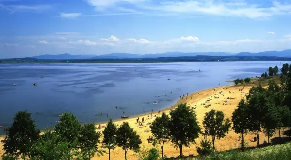 Słoneczna plaża nad Jeziorem Nyskim, pełna ludzi odpoczywających i kąpiących się. W oddali góry, a na wodzie żaglówki i łodzie. To idealne miejsce na wakacyjne atrakcje.