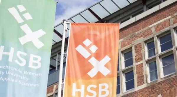 Zwei Fahnen der Hochschule Bremen City University of Applied Sciences wehen vor einem Backsteingebäude.