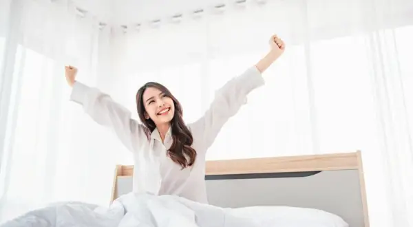 A woman stretches happily in bed, a perfect example of how to wake up with more energy.