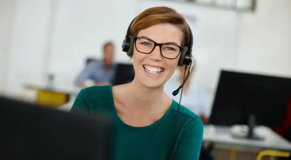 Uśmiechnięta kobieta w okularach i zestawie słuchawkowym, korzystająca z technologii VoIP w biurze.