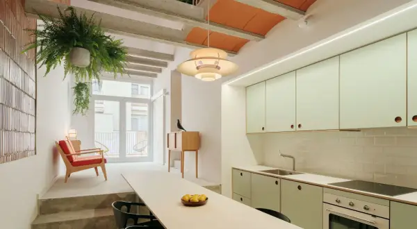 Interior de una casa entre medianeras con cocina moderna, sala de estar con sillón rojo y plantas colgantes.