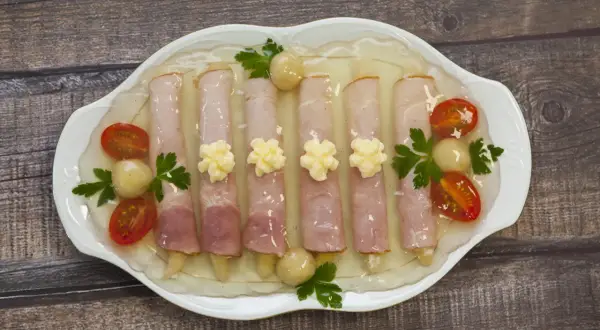 Szparagi w szynce w galarecie, ozdobione pomidorkami koktajlowymi i majonezem, prezentują się apetycznie na białej paterze.