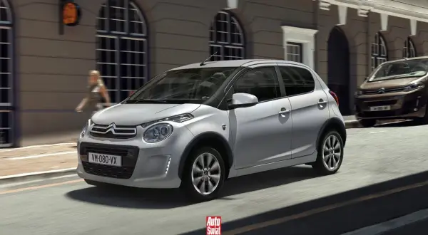 Srebrny Citroën C1 z czarnymi nakładkami na błotniki przemierza miejską ulicę.