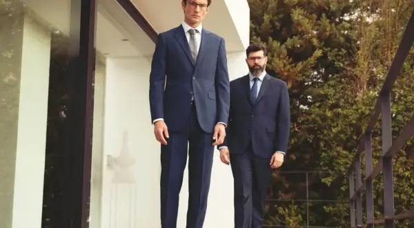 Zwei Männer in eleganten Anzügen stehen auf einer Holzterrasse. Der vordere Mann zeigt, wie man einen Anzug richtig trägt, mit perfekter Passform und Stil.