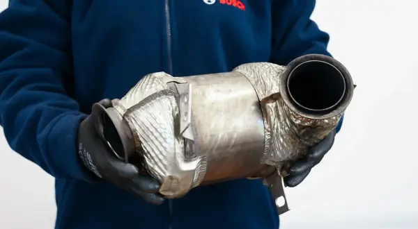 Osoba w rękawiczkach trzyma metalowy element, prawdopodobnie **fap** (filtr cząstek stałych) do samochodu.