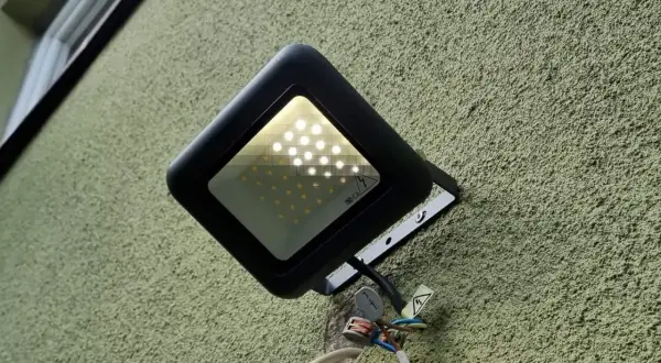 Czarna, nowoczesna lampa naświetlacz LED zamontowana na chropowatej, zielonej ścianie. Widoczne przewody i złączki.