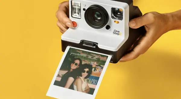 Aparat Polaroid One Step 2 drukuje zdjęcie dwóch kobiet w okularach przeciwsłonecznych na tle kolorowego krajobrazu.