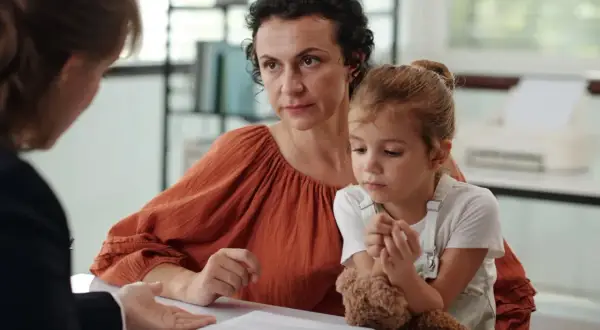 Matka z córką słuchają diagnozy autyzmu. Dziecko tuli misia, a kobieta patrzy z uwagą.