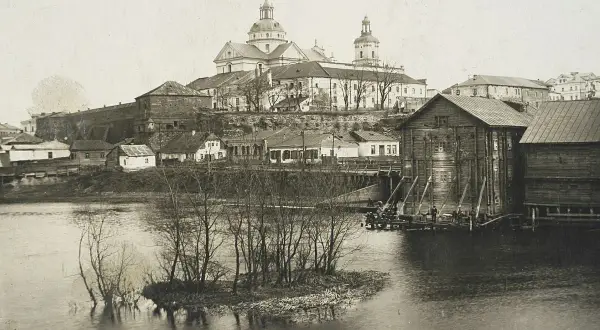 Widok na miasto z rzeką i kościołem w tle. Drewniane budynki nad wodą, mała wysepka z drzewami. Pisz na Berdyczów!