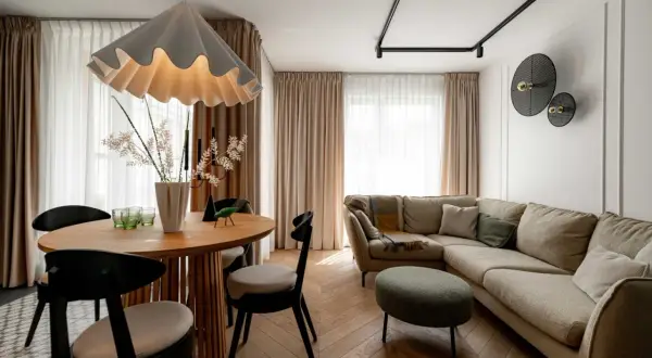 Przytulny salon z beżową kanapą i okrągłym stołem. Nad kanapą w salonie wiszą dekoracyjne lampy, a obok stoi zielona pufa.