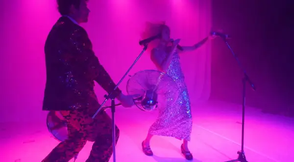 Due cantanti scintillanti, uno con giacca e pantaloni, l'altra con abito, si esibiscono in audizioni musicali sotto luci rosa.