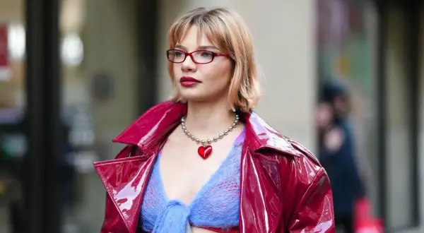 Frau mit roten Brillen und einem stufigen Bob trägt eine glänzende rote Jacke über einem blauen Strickoberteil mit Herz-Halskette.