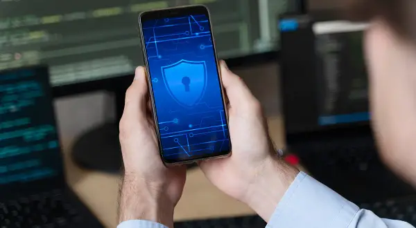 Ręce trzymają smartfon z ikoną tarczy i klucza, symbolizującą bezpieczeństwo. To wskazówka, jak odblokować telefon bez utraty danych.