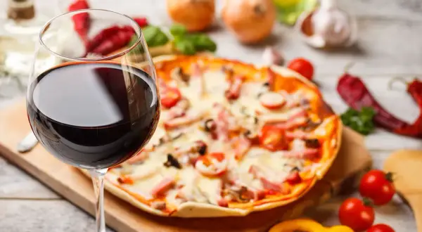 Ein Glas Rotwein neben einer Pizza mit Tomaten und Schinken. Die perfekte Kombination: welcher Wein zu Pizza?