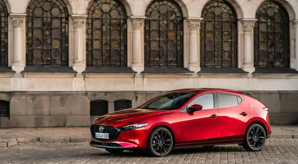 Un elegante Mazda 3 Skyactiv X rojo, con sus líneas fluidas, se detiene frente a una fachada histórica con arcos y vidrieras.