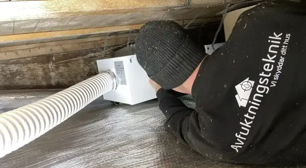 Installering av ventilation i friggebod. En tekniker arbetar med en vit ventilationsenhet under träbjälkar.