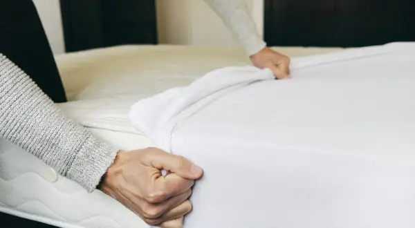Hands pulling a white mattress pad over a mattress. This is part of how to clean a mattress pad.