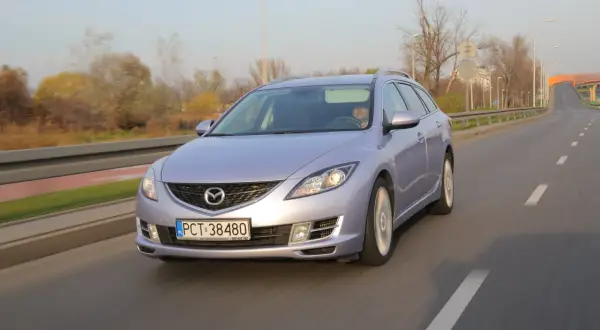 Jasny Mazda 6 kombi jedzie po drodze, jej żarówki mijania oświetlają trasę.
