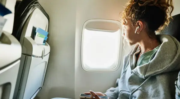 Vrouw met oortjes kijkt uit het vliegtuigraam, hopend op tips wat te doen tegen vliegangst. Ze houdt haar telefoon vast.