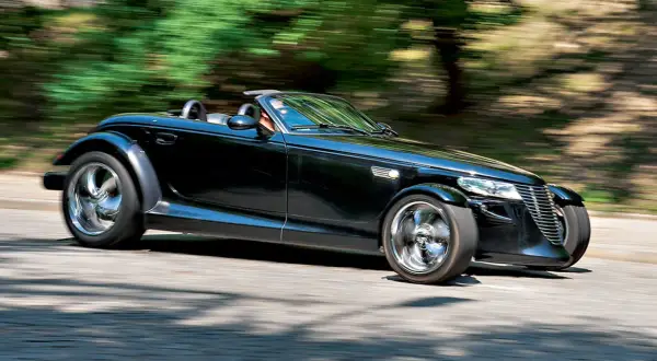 Czarny Plymouth Prowler z opuszczonym dachem pędzi po krętej drodze, otoczony rozmazaną zielenią drzew.