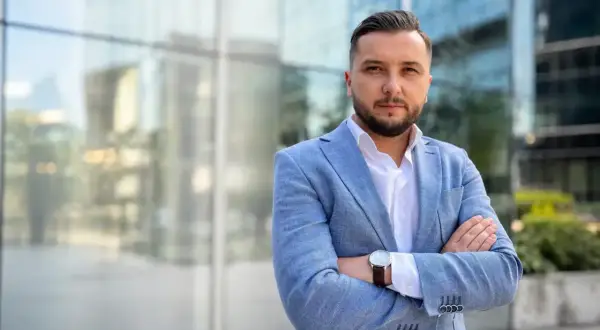 Mężczyzna w jasnoniebieskiej marynarce i białej koszuli, z rękami skrzyżowanymi na piersi, na tle nowoczesnego budynku.