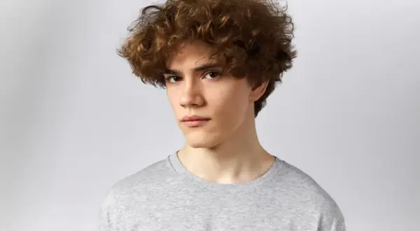 Joven con un peinado rizos hombre, cabello castaño y rizado, viste camiseta gris.