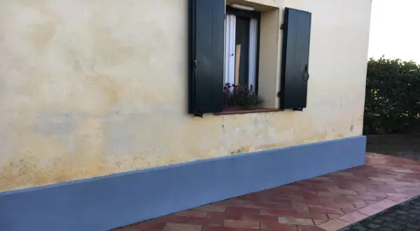 Muro con rinzaffo storico, una finestra con persiane verdi e un davanzale fiorito. Pavimentazione in cotto.