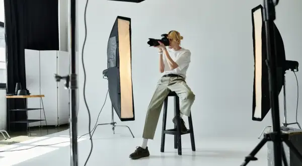 Młody fotograf z blond włosami, siedząc na stołku, robi zdjęcie aparatem. W studiu widać lampy i sprzęt. Zastanawiasz się, ile zarabia fotograf?