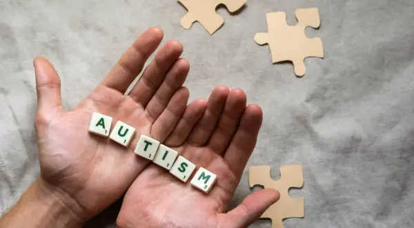 Twee handen houden de letters vast die 'AUTISME' spellen. Puzzelstukjes liggen verspreid, symbolisch voor het spectrum.