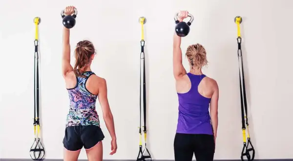 Dwie kobiety wykonują ćwiczenia na ból barku z kettlebellami. W tle wiszą taśmy TRX.