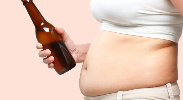 Dłoń trzyma butelkę piwa, obok widoczny brzuch piwny.