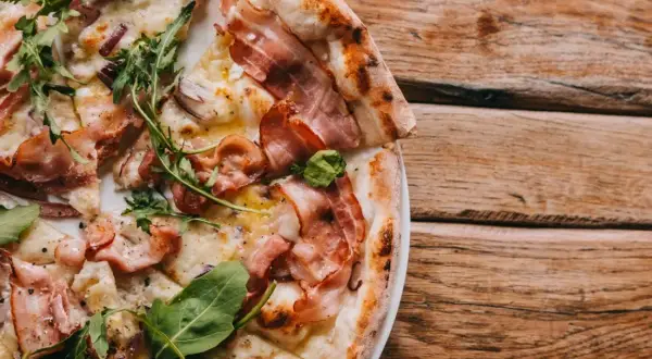 Pyszna pizza z chrupiącym rozczynem na pizze, boczkiem i rukolą, podana na drewnianym stole.