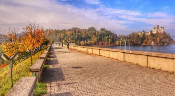 Jesioro Czorsztyńskie jesienią. Spacerowicze i rowerzyści na promenadzie, w tle zamek i kolorowe drzewa.