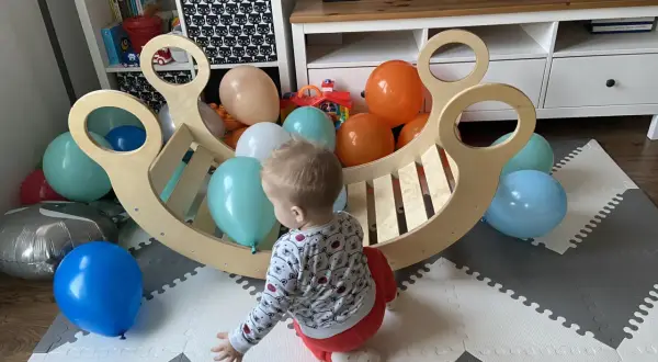 Roczne dziecko bawi się z kolorowymi balonami przy drewnianej kołysce. To idealne miejsce do rozwoju metodą Montessori.