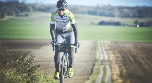 Kolarz w stroju sportowym jedzie rowerem po szutrowej drodze wśród pól.