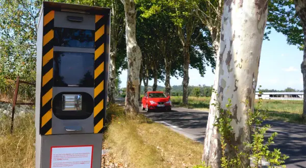 Panneau de **détecteur de radar interdit** sur le bord de la route, avec une voiture rouge passant à proximité.