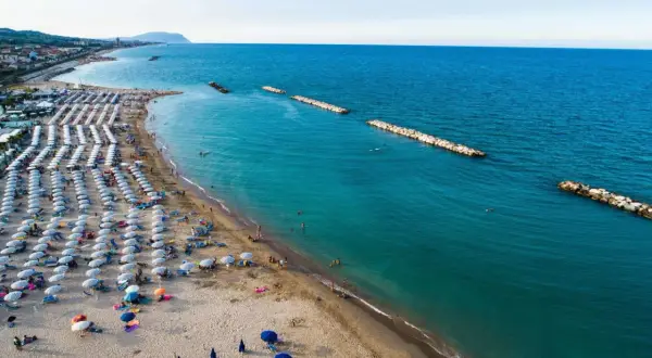 Ampia spiaggia libera a Civitanova Marche, con file ordinate di ombrelloni e lettini, bagnanti che si godono il mare turchese e dighe frangiflutti in pietra.