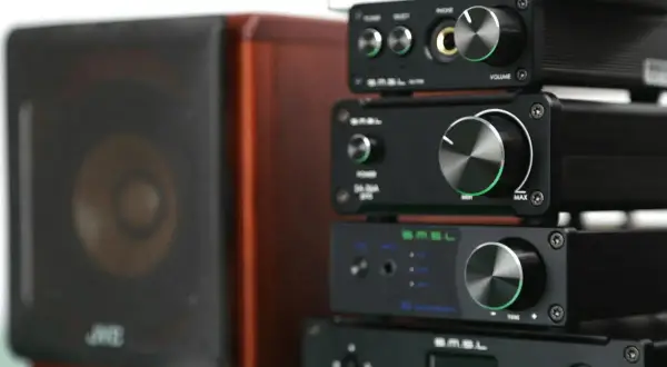 Stosik czarnych urządzeń audio SMSL, w tym SMSL M6 Mini DAC, obok brązowej kolumny głośnikowej.