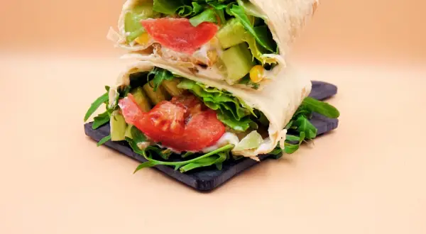 Dietetyczna tortilla z warzywami: sałatą, pomidorem, ogórkiem i rukolą, na ciemnej desce.