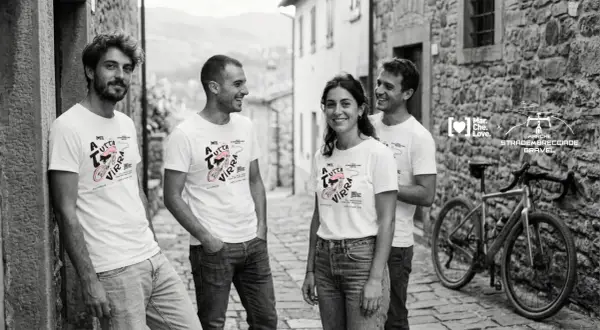Quattro amici sorridono in un vicolo di pietra, indossando magliette con un disegno di bici e la scritta "A tutta virra". Un'espressione del vocabolario dialetto marchigiano che evoca passione.
