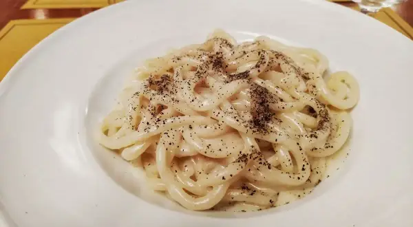 Piatto di pici cacio e pepe, pasta fresca condita con pecorino romano e pepe nero macinato fresco, servita su un piatto bianco.
