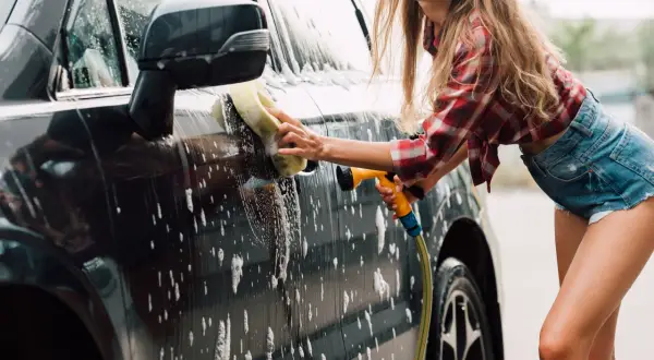 Uśmiechnięta kobieta myje auto gąbką i wężem. Domowe sposoby na czysty samochód to świetna zabawa!